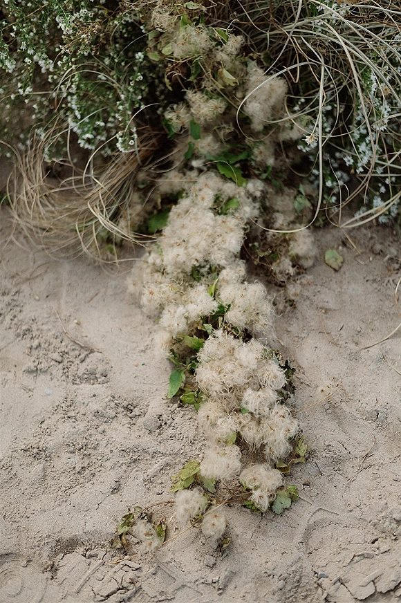 Aisle floral decor with a ground floral arrangement of white wildflowers and trailing greenery on sand, with natural greenery behind