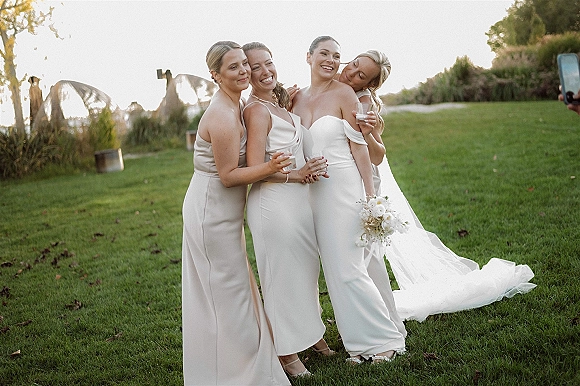 Bridesmaid photo of bride with bridesmaids hugging her, holding champagne coupe glasses on a garden lawn at sunset with veil and train