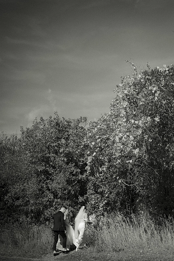 Couple portrait of bride and groom walking away on a field path, her veil blowing and bouquet in hand, framed by tall grass and trees