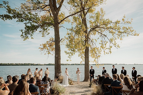 Ceremony moment at an outdoor wedding ceremony with bride in veil and groom in tux at the lakeside altar, framed by trees and guests seated