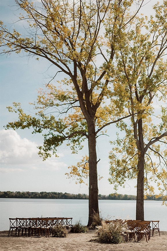 Ceremony setup with outdoor ceremony seating of wood cross back chairs lined down a sandy lakeside aisle with pampas grass accents under tall trees