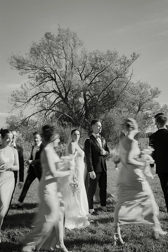 Wedding party portrait of bride and groom with wedding party, bride in veil holding bouquet with ribbon as bridesmaids walk by on a lawn