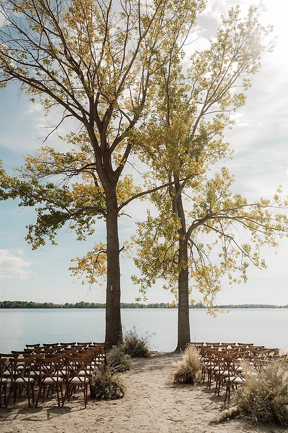 Ceremony setup with wooden crossback chairs and grounded aisle florals on sandy lakeside shoreline, framed by tall trees and water view