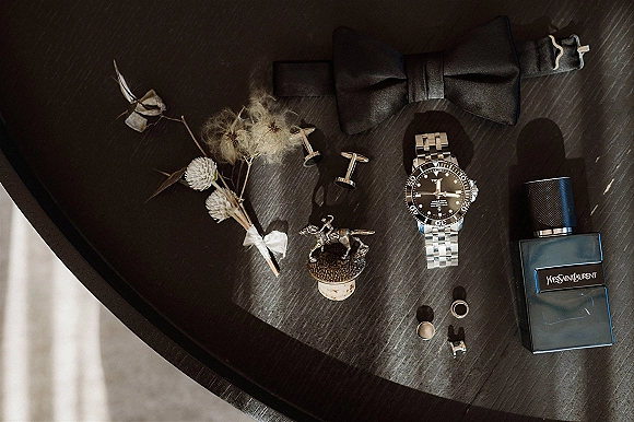 Groom accessories flatlay with black bow tie, watch, cufflinks, boutonniere, cologne, rings, and a small figurine on dark wood tabletop