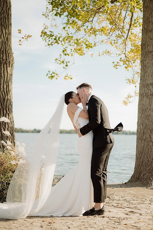 Wedding kiss as bride in strapless dress and long veil embraces groom in black tuxedo on a sandy lakeside shore under trees