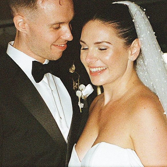 Wedding couple portrait, bride and groom close up as she smiles in a strapless gown while he looks at her in a tuxedo against dark indoors