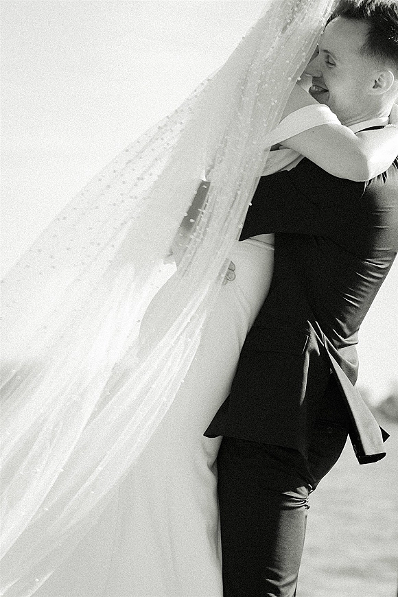 Wedding kiss portrait of bride and groom kiss under a long veil, side profile embrace in classic attire against a bright sky