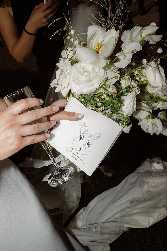 Wedding bouquet of white flowers and greenery with ribbon, held beside a champagne flute and illustrated place card in dim reception light