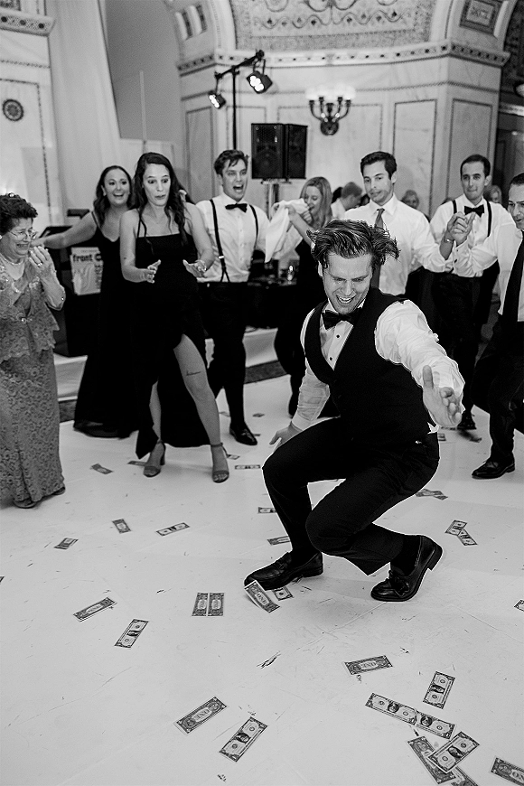 Wedding reception dancing with groom in bow tie and vest leading a money dance as guests cheer under stage lights in a ballroom