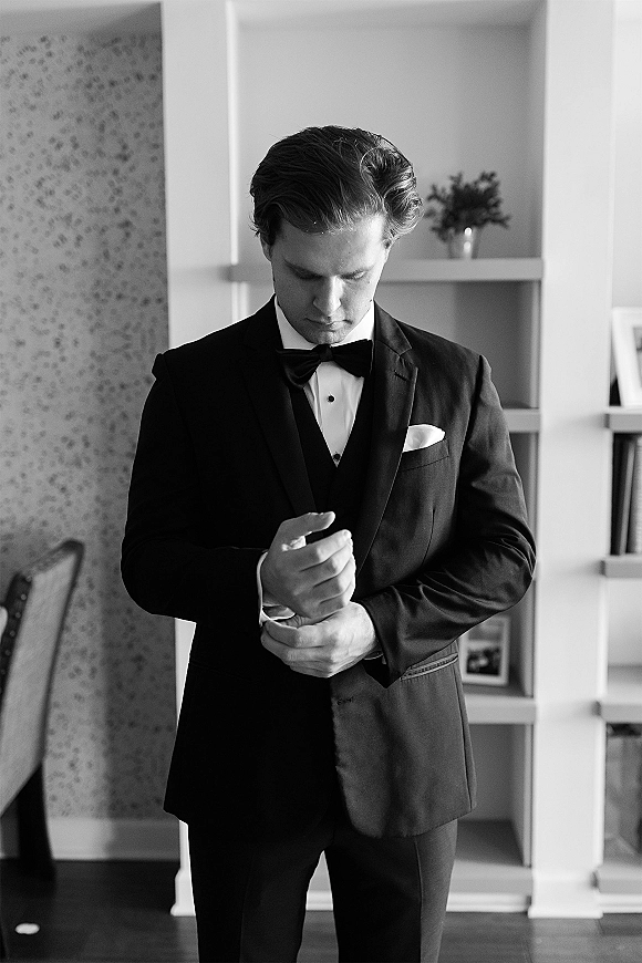 Groom portrait in a black tuxedo adjusting cufflinks, bow tie and boutonniere, standing by a bookshelf with floral wallpaper indoors