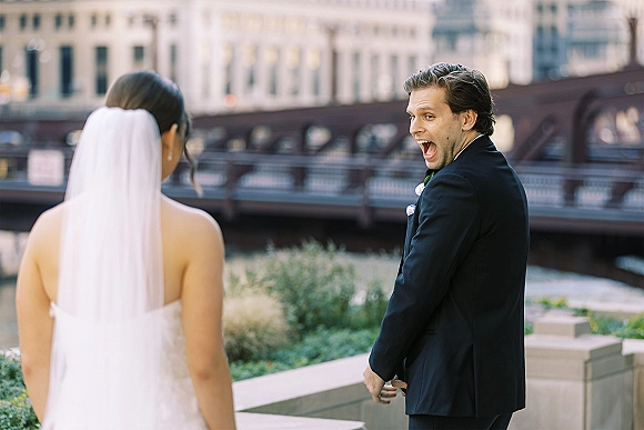 First look moment as bride in veil and strapless gown walks up behind groom in black suit on a riverwalk by a bridge and city skyline