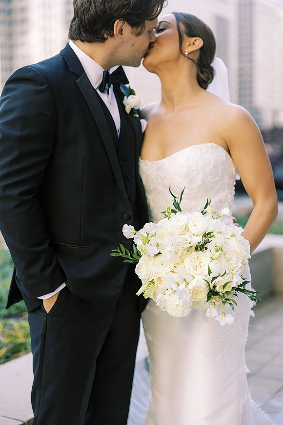 Wedding kiss portrait of bride and groom kissing, her strapless lace dress and veil beside his black tuxedo on a city walkway with greenery