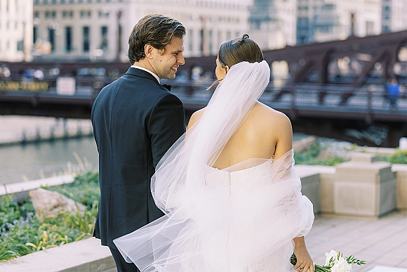 Couple portrait of bride and groom walking by a riverfront, bride looking back with long veil and calla lily bouquet, city bridge behind
