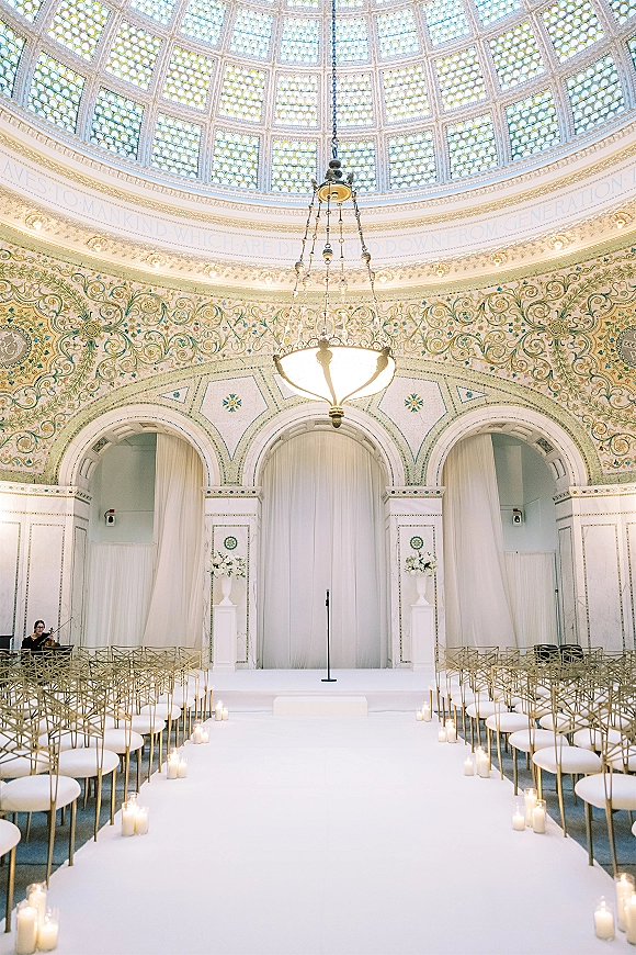 Ceremony setup with a white aisle runner lined by pillar candles, gold chairs and draped altar beneath a stained glass dome ceiling