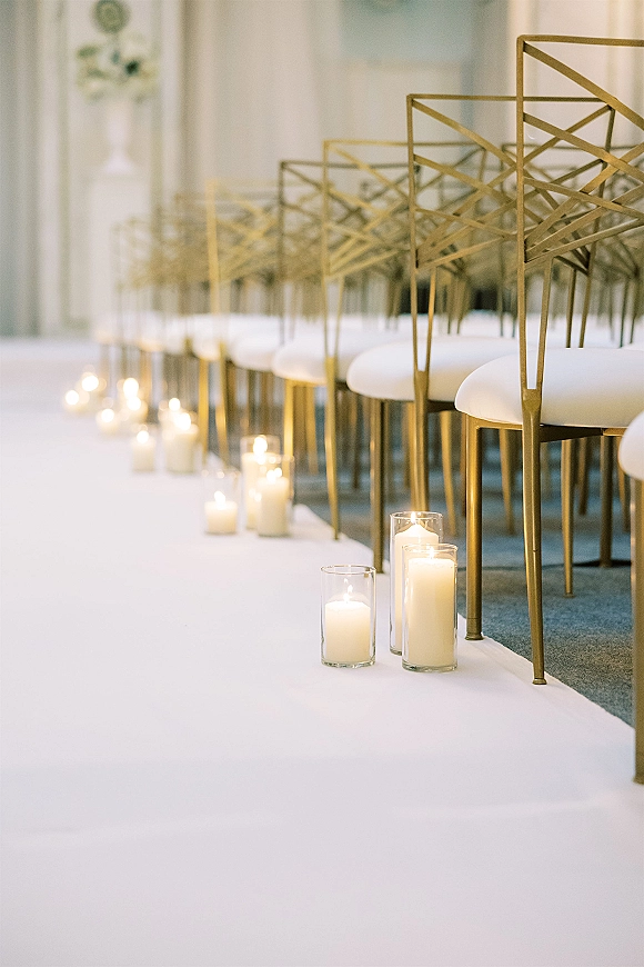 Ceremony aisle decor with wedding aisle candles in glass cylinder and pillar styles lining a white runner beside gold chairs in an indoor venue