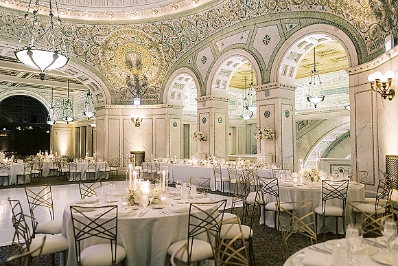 Reception ballroom decor with luxury wedding reception tables, white linens, gold chairs, candlelit floral centerpieces beneath arched ceilings and chandeliers