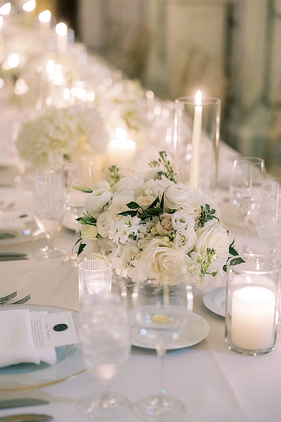 Reception tablescape with a wedding table centerpiece of white roses and greenery, taper and hurricane candles, place settings, and soft bokeh lights