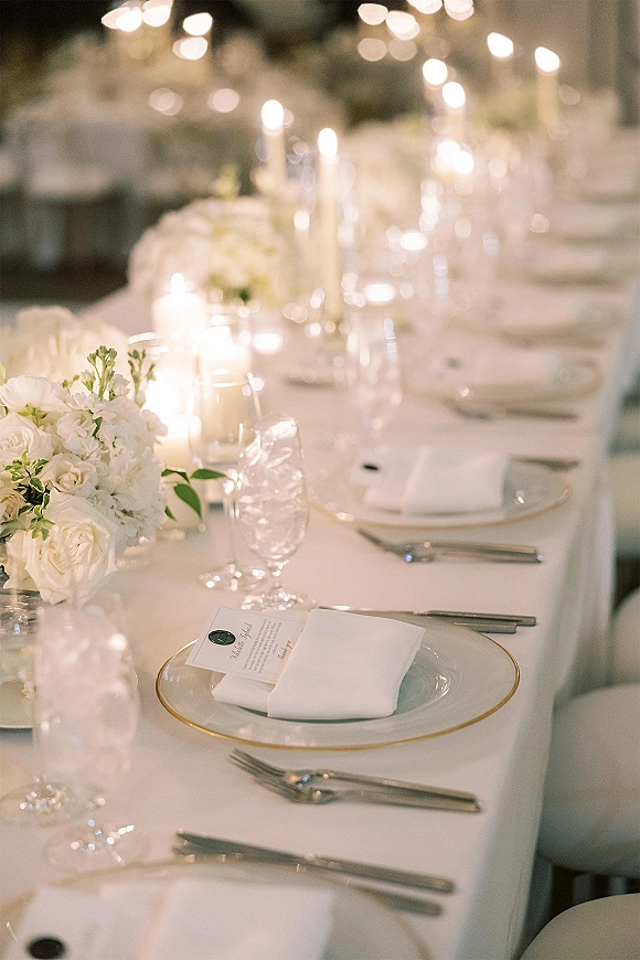 Reception tablescape with wedding reception table decor featuring white floral centerpieces and taper candles along a long banquet table under soft lighting