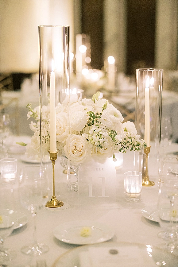 Reception tablescape with a white floral centerpiece in tall hurricane vases, taper candles, gold candlesticks, and place settings in a candlelit banquet hall