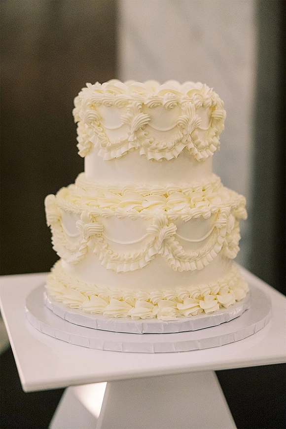 Wedding cake with white buttercream frosting, two-tier design and piped ruffle scalloped icing on a cake stand against a neutral wall