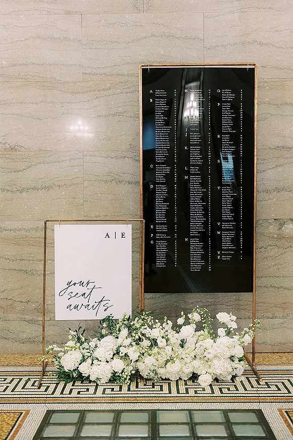 Wedding seating chart acrylic seating chart in a gold frame stand with calligraphy, white rose florals and greenery against a stone wall