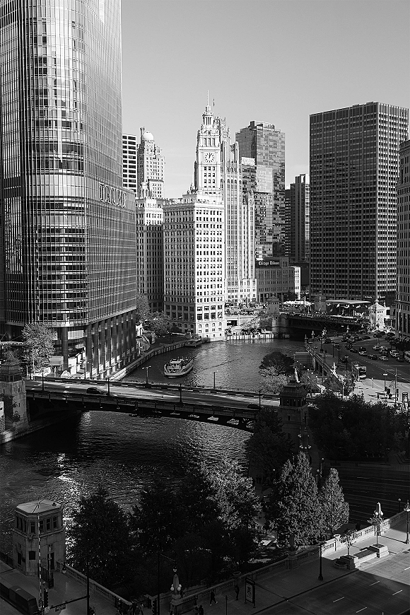City skyline photo in black and white cityscape with a river, bridge, and boat, framed by skyscrapers and a clock tower above streets
