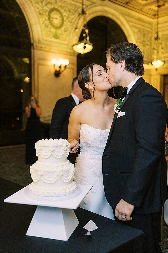 Wedding cake cutting as the bride and groom kiss beside a two-tier white cake on a stand in a chandelier-lit ballroom with guests