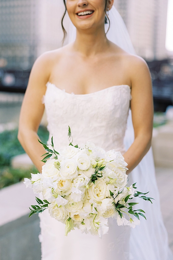 Bridal portrait of a bride holding bouquet, smiling in a strapless lace wedding dress with veil and earrings by a stone facade walkway