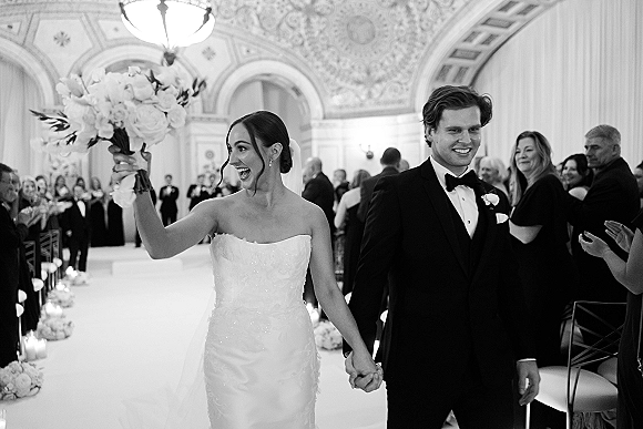 Wedding recessional as bride and groom walk the aisle holding hands, bride lifting bouquet, candlelit hall with chandelier and guests cheering