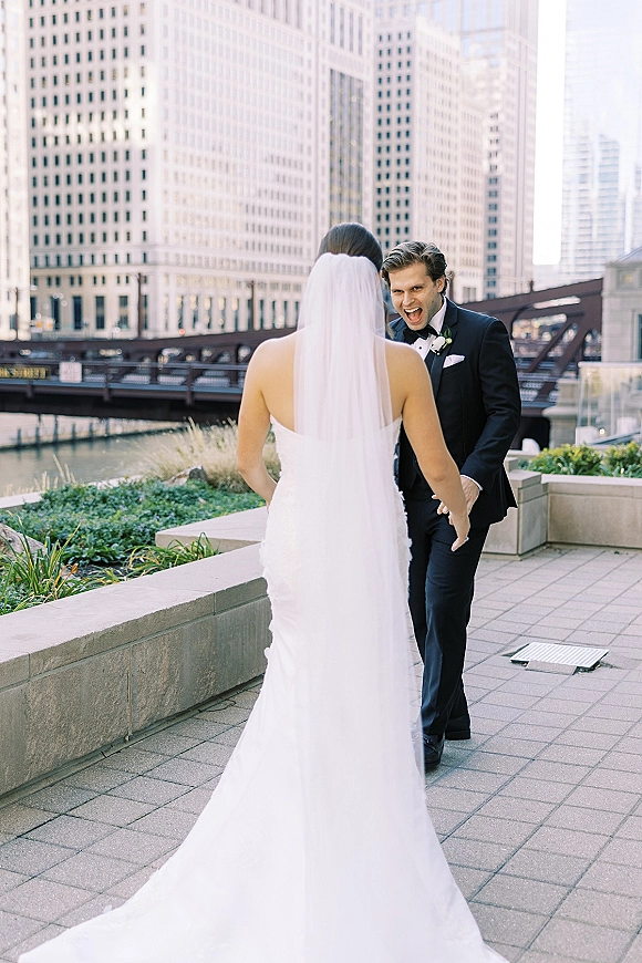 First look moment as bride in a long veil walks up behind groom in black tuxedo on a riverwalk with bridge and city buildings behind