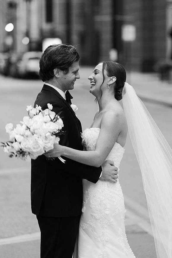 Couple portrait of bride and groom laughing as they embrace, bride holding a rose bouquet and veil flowing on a city street backdrop