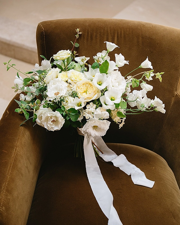 Bridal bouquet with white rose blooms and sweet peas, wrapped in flowing white ribbon, resting on a brown velvet chair against a neutral wall