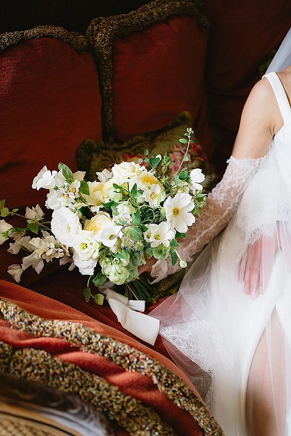 Bridal bouquet with white and green bouquet blooms, greenery, and ribbon beside a bridal veil and lace sleeve on a vintage chair