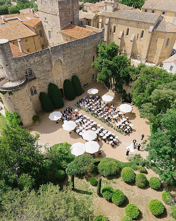 Outdoor wedding reception in a castle courtyard with round tables, white patio umbrellas, and greenery garlands beside stone walls