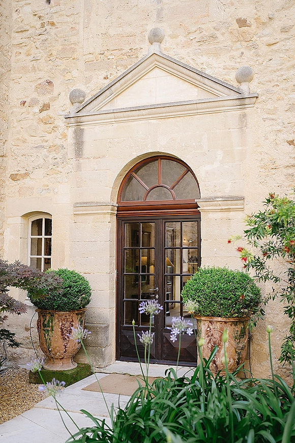 Wedding venue exterior with glass double doors under an arched transom, framed by potted topiary along a gravel entry walkway