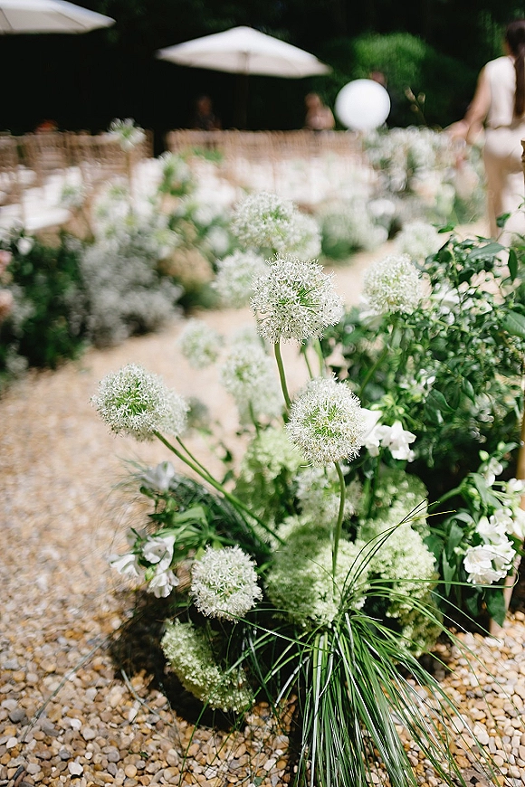 Ceremony aisle flowers with white allium and greenery accents lining a gravel path between garden chairs under white umbrellas