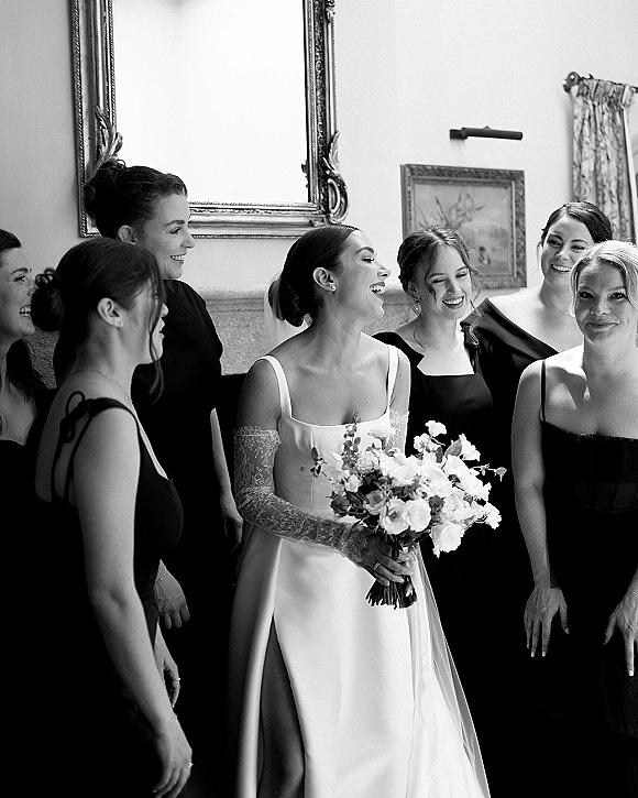 Bride with bridesmaids getting ready, laughing in a hotel room as she holds a white bouquet, wearing lace gloves and satin gown