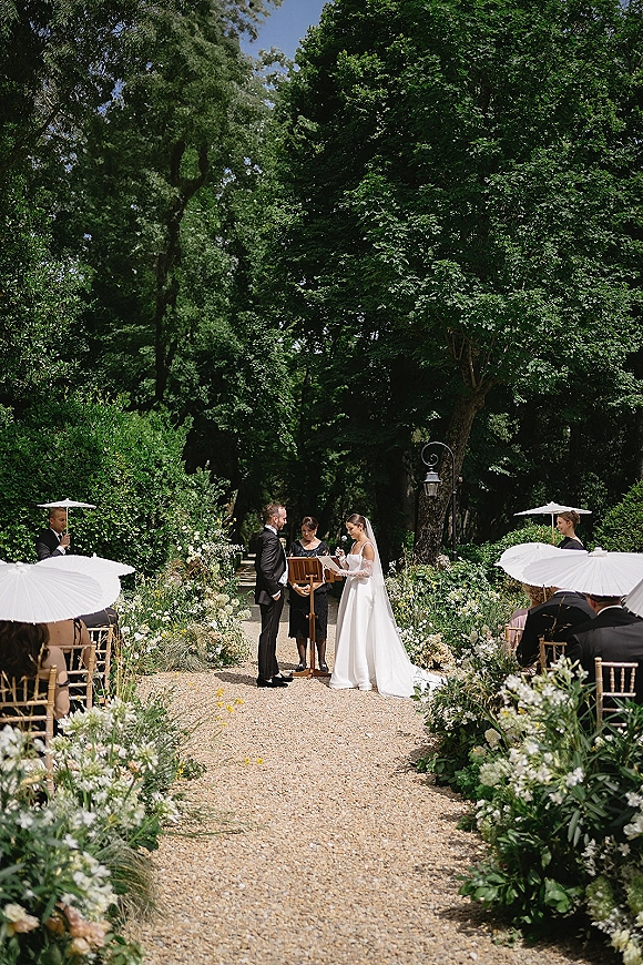 Wedding vows as the bride reads into a handheld microphone beside the groom at a wooden lectern, veil flowing on a flower-lined garden aisle