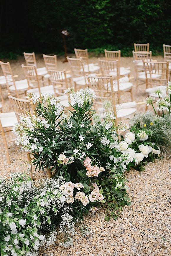 Ceremony aisle florals with grounded aisle flowers in white, blush, and greenery lining a gravel path beside chiavari chairs in a garden setting