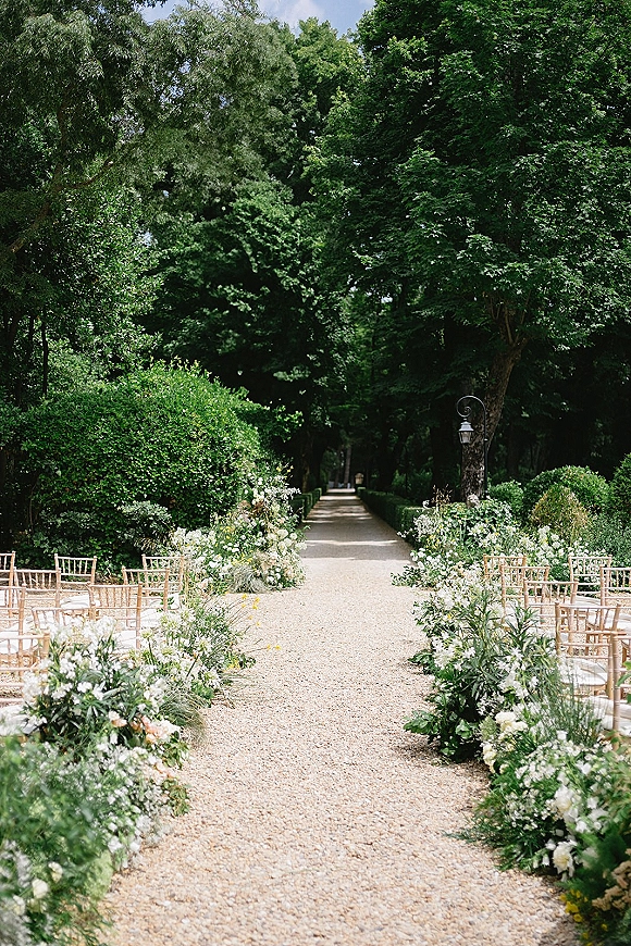 Ceremony aisle design with white and green meadow florals lining a gravel walkway, wooden Chiavari chairs, and a lantern on a tree-lined path