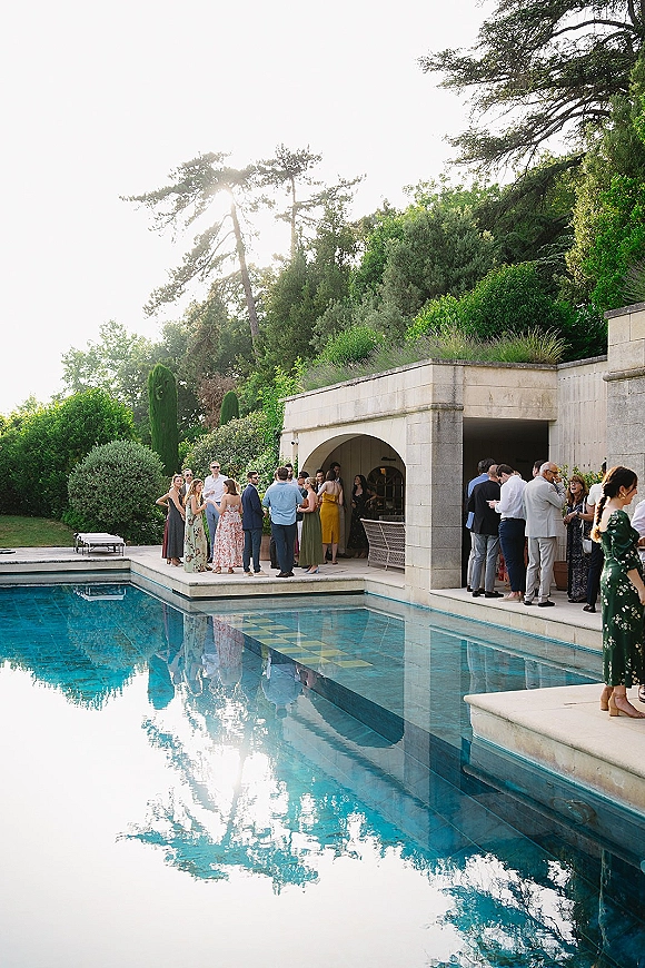 Wedding cocktail hour by the pool with outdoor wedding reception drinks at a bar counter, lounge chairs on a stone terrace by an arched pavilion