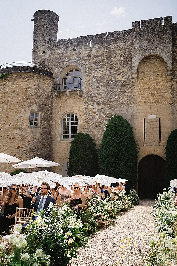 Outdoor ceremony setup with a floral lined aisle and wooden chairs beneath white parasols in a stone castle courtyard by an arched doorway