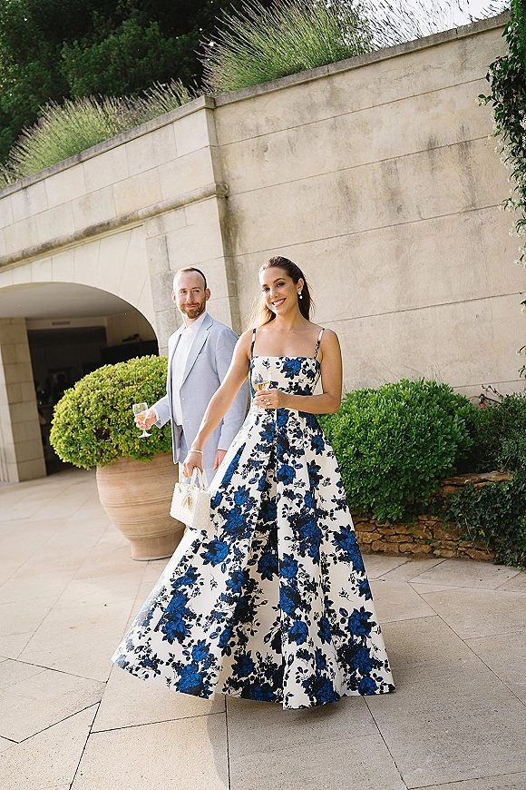 Wedding guest outfit in a blue floral wedding guest dress holding a white handbag and champagne beside a light suit by a stone archway