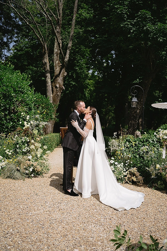 Wedding kiss portrait of bride and groom kissing, her long veil and lace sleeves flowing on a gravel garden path by a vintage lamppost