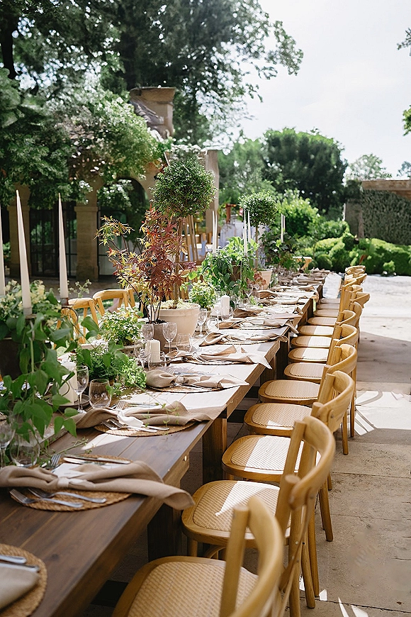 Reception tablescape on a long wooden banquet table with beige napkins, woven placemats, taper candles and greenery on a stone patio