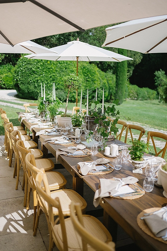 Reception tablescape on a wood farmhouse table with woven placemats, beige napkins, taper candles, greenery, and white patio umbrellas on a garden lawn