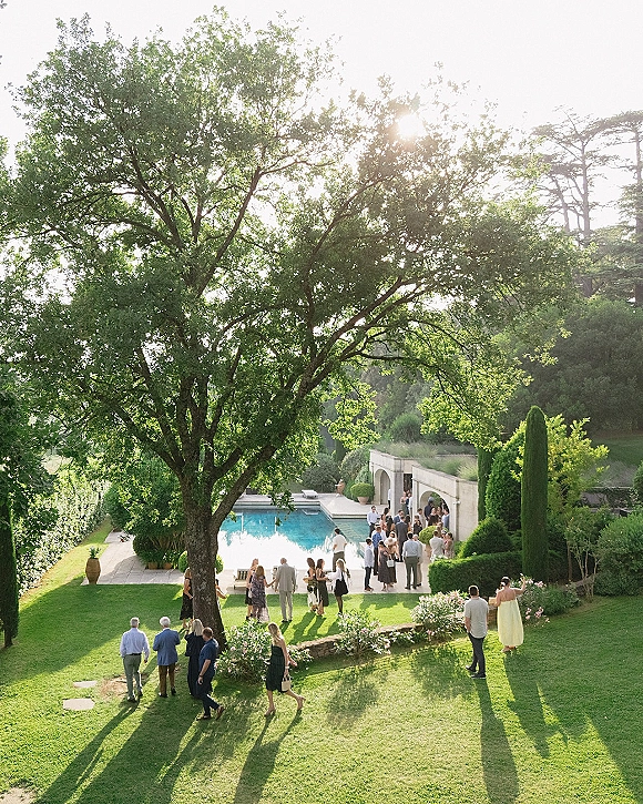 Wedding cocktail hour with outdoor wedding cocktail hour guests mingling by a swimming pool on a stone patio with garden planters and sun flare