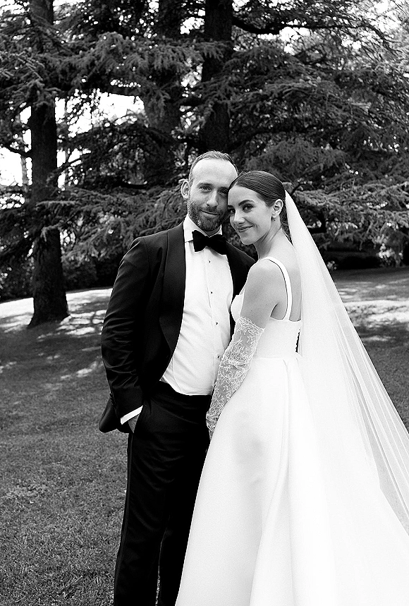 Couple portrait in a close embrace, bride in veil with lace gloves and groom in tuxedo with bow tie on a lawn by evergreens
