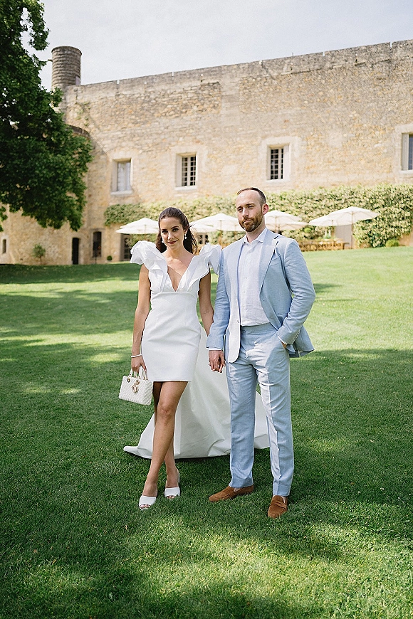 Couple portrait of newlyweds holding hands, bride in a short wedding dress with ruffle sleeves beside groom in a light blue suit by an ivy wall