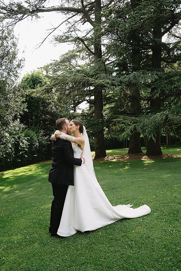 Wedding kiss portrait of bride and groom kissing on a green lawn, her long veil and lace-sleeve dress train flowing by evergreen trees